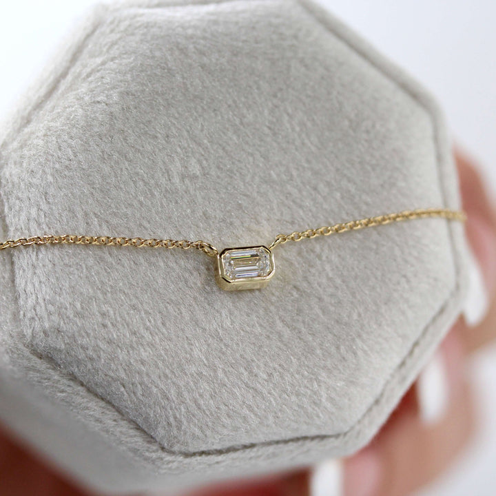 The East-West Emerald Bezel Necklace in Yellow Gold against a gray velvet ring box held by a hand