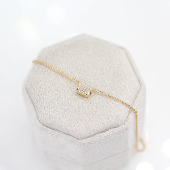 The East-West Emerald Bezel Necklace in Yellow Gold atop of a gray velvet ring box