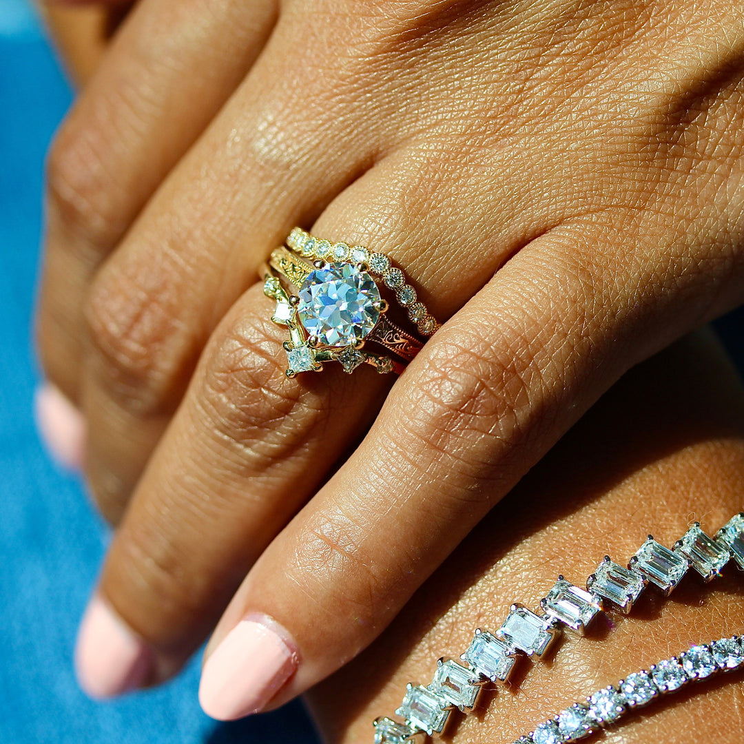 The Kathleen ring in yellow gold stacked with the Florence Contour wedding band in yellow gold and the Astra wedding band in yellow gold modeled on a hand with the Emerald Lab Grown Diamond Tennis Bracelet in white gold and the Classic 6.02ct Lab Grown Diamond Tennis Bracelet in white gold