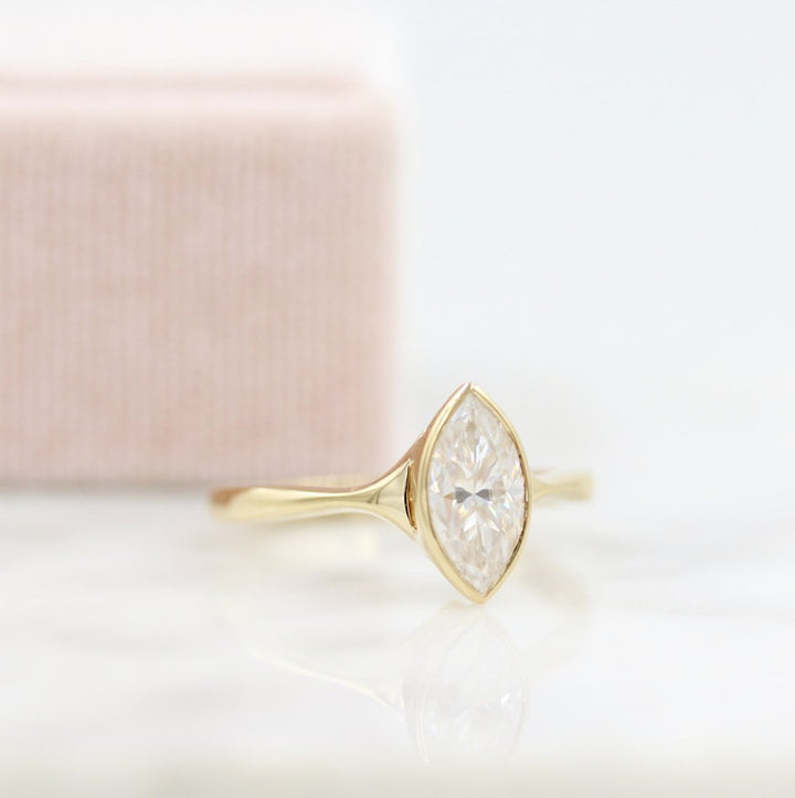 Yellow gold bezel solitaire with marquise moissanite center and light pink velvet ring box in the background