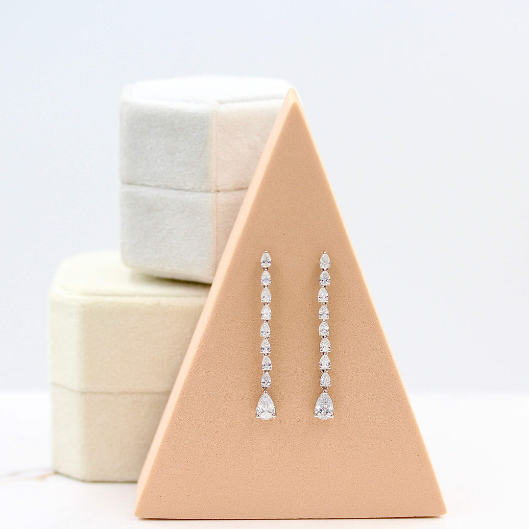 Pear Drop Earrings in white gold hung on a salmon foam triangle-shaped block in front of two stacked ring boxes