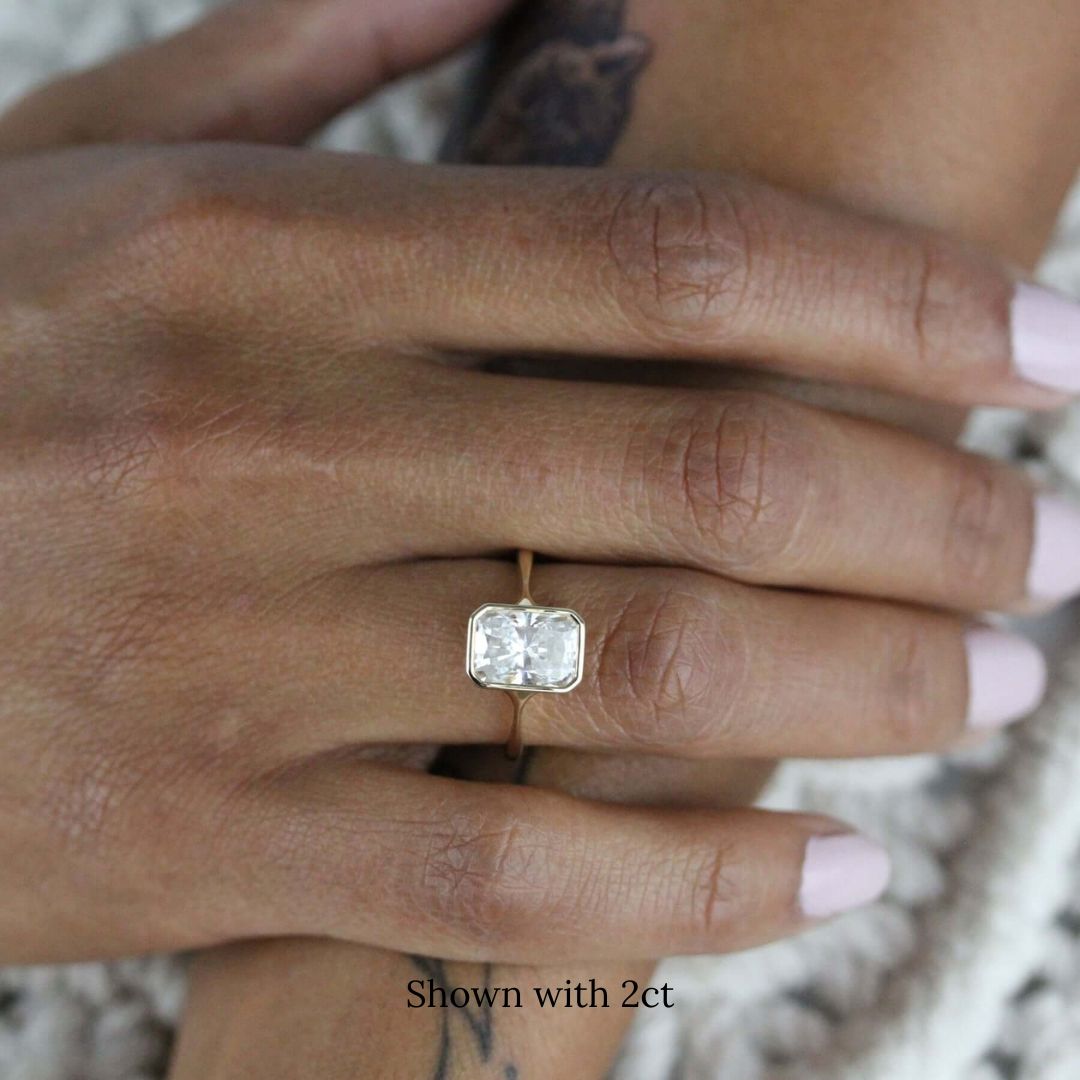 Hand wearing 2ct radiant moissanite solitaire engagement ring in rose gold, resting on tattooed arm