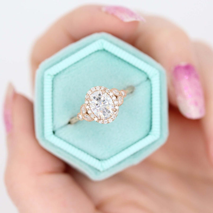 The Cate Ring (Oval) in rose gold in a blue velvet ring box held by a hand with pink fingernails