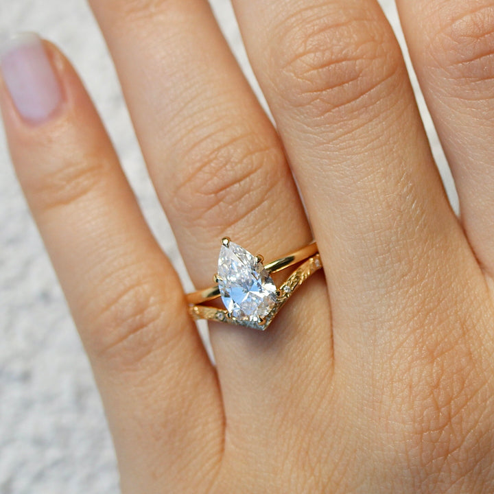 Yellow Gold V shaped Willow Wedding Band with a Pear-shaped Diamond Engagement Ring on a hand against a neutral background.