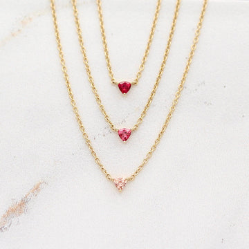 The July Tiny Trillion Birthstone Necklace with Ruby in Yellow Gold stacked with the Janurary Tiny Trillion Birthstone Necklace with Padparadscha Sapphire in Yellow Gold and the October Tiny Trillion Birthstone Necklace with Peachy-Pink Sapphire in Yellow Gold against a white background