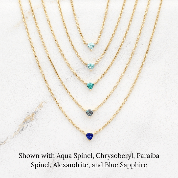 The March Tiny Trillion Birthstone Necklace with Aqua Spinel in Yellow Gold stacked with the August Tiny Trillion Birthstone Necklace with Chrysoberyl in Yellow Gold, the December Tiny Trillion Birthstone Necklace with Paraiba Spinel in Yellow Gold, and the June Tiny Trillion Birthstone Necklace with Alexandrite, and the September Tiny Trillion Birthstone Necklace with Blue Sapphire in Yellow Gold in Yellow Gold against a white background