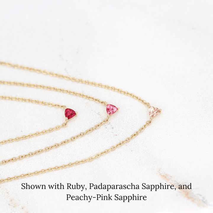 The July Tiny Trillion Birthstone Necklace with Ruby in Yellow Gold stacked with the January Tiny Trillion Birthstone Necklace with Padparadscha Sapphire in Yellow Gold and the October Tiny Trillion Birthstone Necklace with Peachy-Pink Sapphire in Yellow Gold against a white background