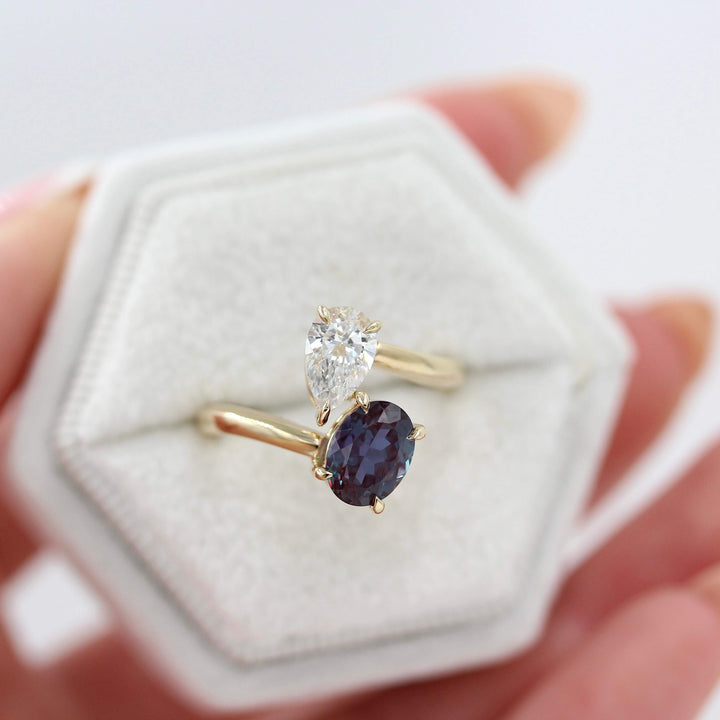 Toi et Moi Diamond and Alexandrite Bypass Ring in yellow gold against a white background in a white velvet ring box held by a hand