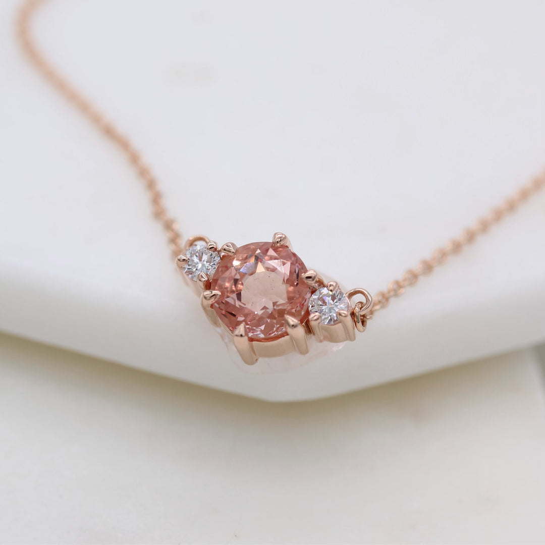 rose gold necklace with peachy-pink sapphire center and round diamond accents on a rose gold chain with a white background